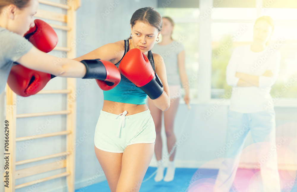 Concentrated adolescent girl in boxing gloves practicing punches in sparring during group self defence course supervised by female instructor in gym..
