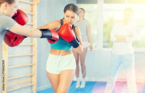 Concentrated adolescent girl in boxing gloves practicing punches in sparring during group self defence course supervised by female instructor in gym.. © JackF