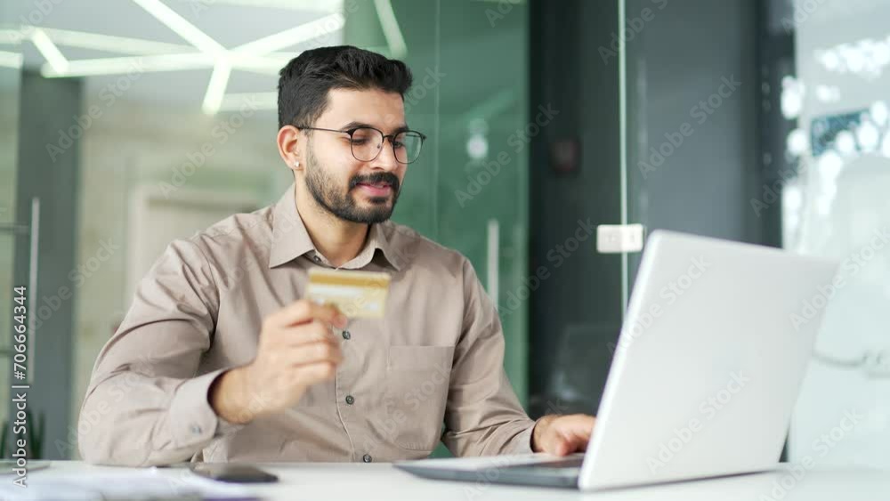 Smiling bearded entrepreneur enters a credit card number on a laptop while sitting at a desk at a workplace in business office. Handsome businessman in glasses shopping online, happy about good price