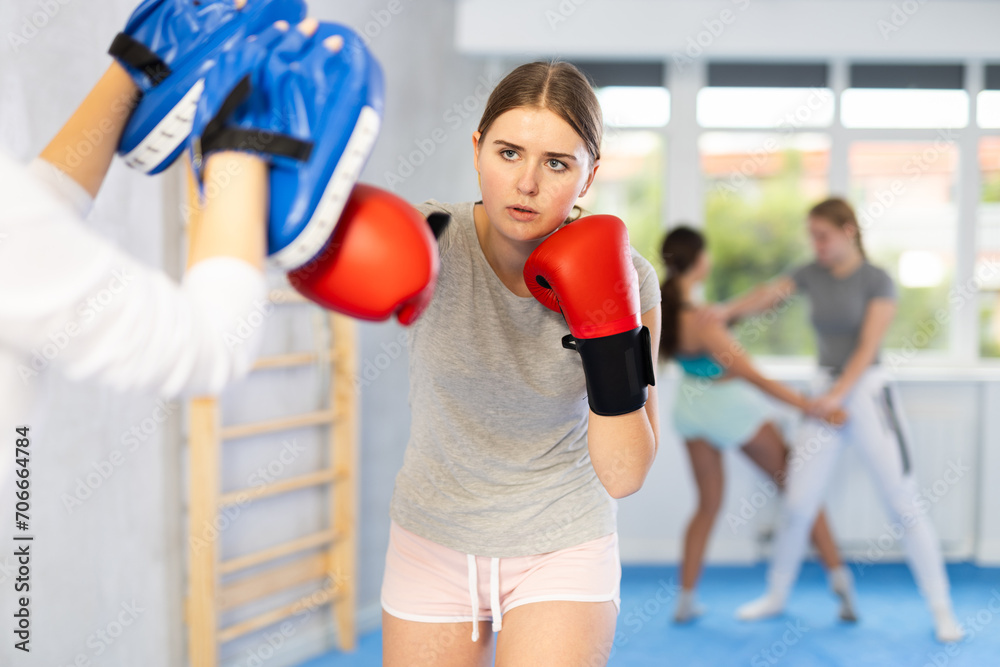 Focused teenage girl wearing boxing gloves working out with female coach during self-defense course, practicing punches on mitts..