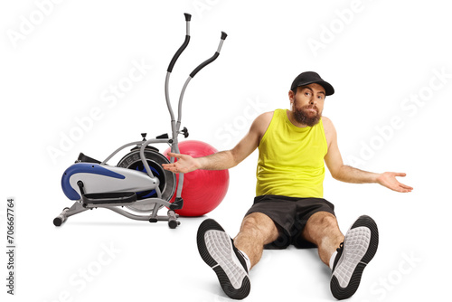 Confused bearded man in sportswear sitting on the floor in front of gym equipment