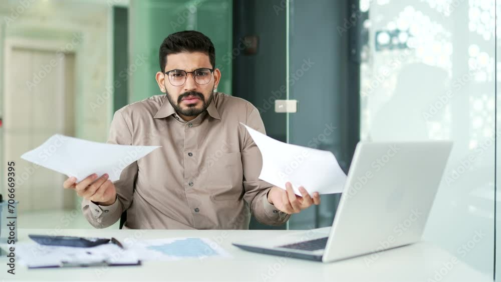 Confused businessman in glasses having difficulty with paper work sitting at workplace in business office. Puzzled entrepreneur looks at documents and cannot understand the problem looking at camera