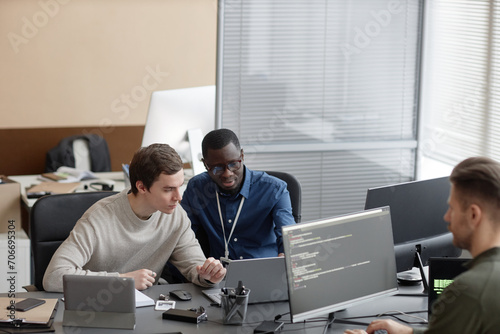 Wide angle shot of African American IT male mentor pointing out inaccuracies in code of young Caucasian male programmer while sitting with laptop at desk photo
