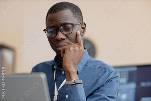 Medium close up shot of thoughtful African American male IT company employee wearing glasses working on laptop at table