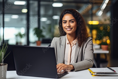 Smiling Young Latina Business Professional Using Laptop at Office Desk, Contemplating Corporate AI Technology Solutions