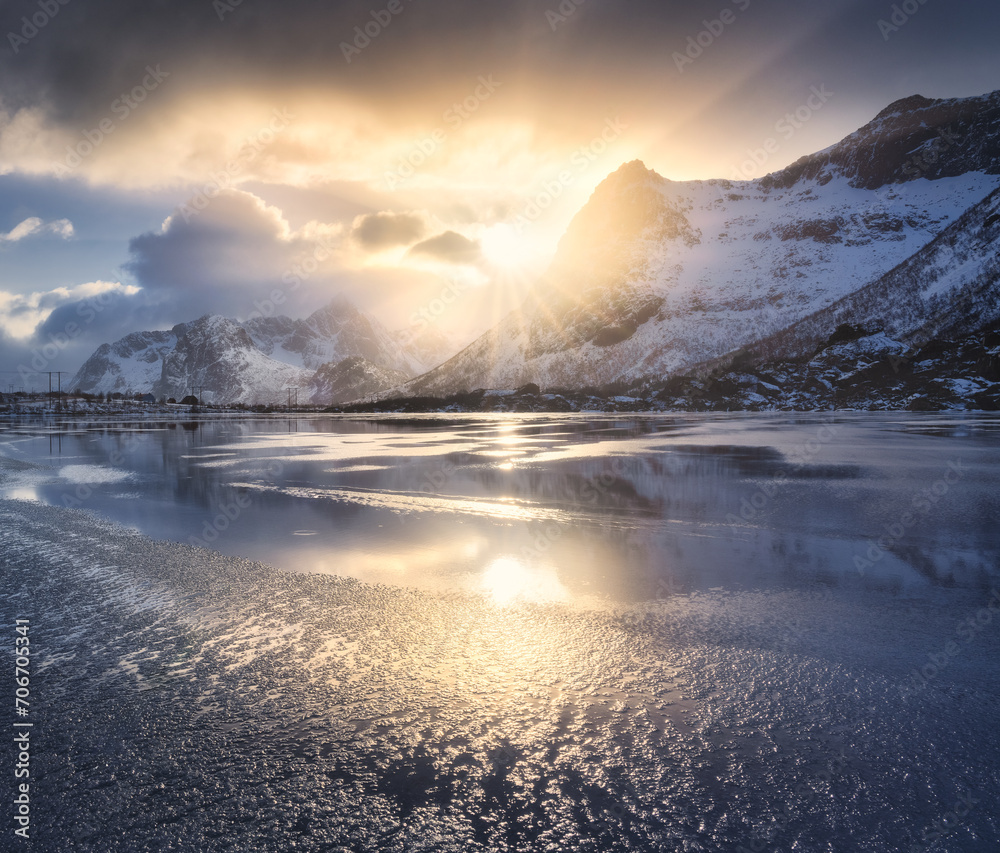 Beautiful snowy mountains and colorful sky with clouds and golden sunlight at sunrise in winter in Lofoten islands, Norway. Landscape with rocks in snow, frozen sea coast, reflection in water. Nature