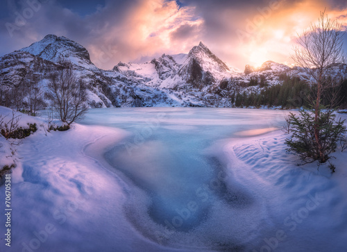 Frozen sea coast and rocks in snow at colorful sunset. Winter in Lofoten islands, Norway. Snowy mountain peaks, lake with frosty shore, ice, trees, colorful sky with clouds golden light. Landscape © den-belitsky