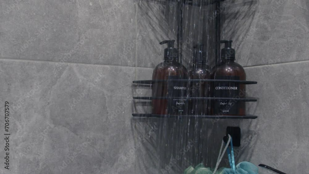 Water flows from the shower in slow motion against the background of gray tiles and bottles of shampoo and conditioner standing on the shelf. Water flows from the shower in slow motion against the background of gray tiles and bottles of shampoo and conditioner standing on the shelf.