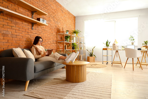 Young woman relaxing on sofa reading book in modern living room with brick wa...
