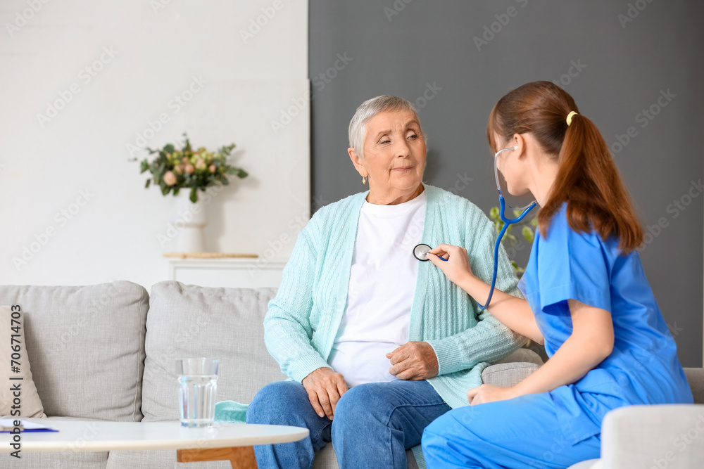 Fototapeta premium Young caregiver hearing senior woman with stethoscope at home
