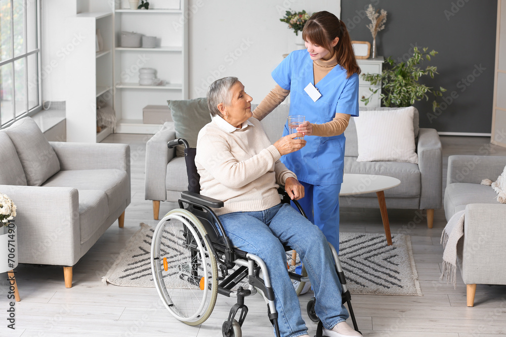 Senior woman in wheelchair taking glass of water from caregiver at home Senior woman in wheelchair taking glass of water from caregiver at home