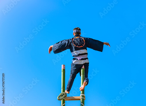 A Japanese hikeshi firefighter standing on one leg at the top of a tall bamboo ladder performing acrobatics during the traditional Hashigo-nori show of Suga shrine against the blue sky of Tokyo.
