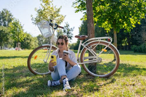 Smiling young hipster woman with bicycle using mobile phone in city park