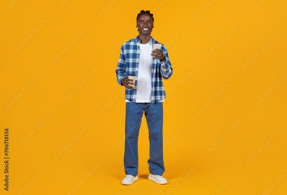 Fototapeta premium Smiling young black man holding takeaway coffee and smartphone