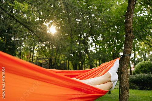Legs of a woman on an orange hammock against the background of green trees and sunlight. The concept of recreation and fulfillment in nature, hiking, escape from the city, festival and vacation