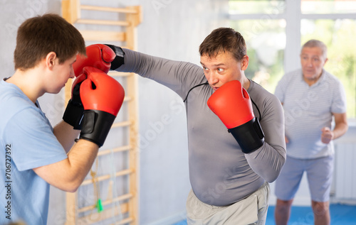Focused man in boxing gloves working out under guidance of experienced instructor during self-defense course, practicing basic punches in sparring with guy