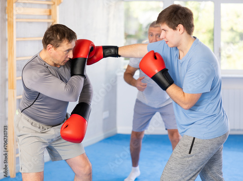 Determined young guy mastering self-defense techniques, practicing punches in sparring with adult man under guidance of experienced coach at boxing gym.. © JackF