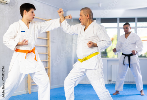 Determined motivated senior man wearing kimono working on hand strikes and martial arts skills in sparring with young opponent during group training