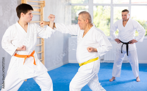 Experienced elderly karate fighter in kimono with yellow belt engaging kumite with young rival, demonstrating martial art techniques in training room