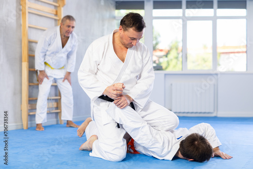 Focused adult jiu-jitsu fighter performing painful armlock on male opponent lying on tatami during sparring in training room