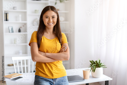 Portrait Of Smiling Young Asian Woman Standing With Folded Arms Near Desk
