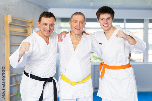 Three generations of men, older, middle-aged, and young, dressed in kimonos friendly embracing in training room. Concept of unity and traditions of family of martial arts fighters..