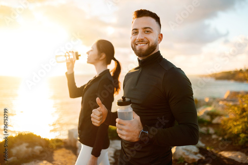 Active couple on coastal trail, man with water bottle giving thumbs up, woman drinking in background photo