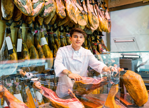 Young male seller cutting slices from whole leg of jamon in meat store