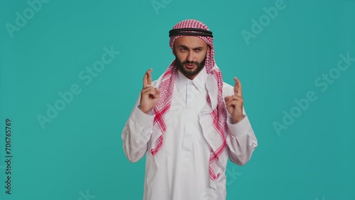 Arab person poses with fingers crossed and praying to allah over blank background, showcasing religion and spiritual devotion. Man in muslim clothes being religious, worshipping God with faith. photo