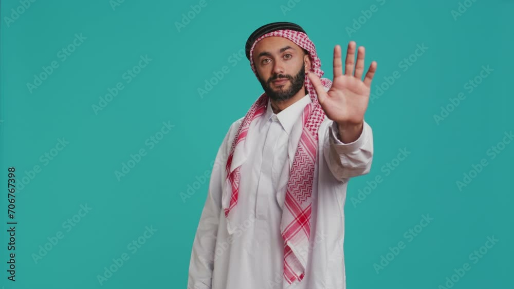 Muslim man showing stop symbol with palm, presenting negativity and disapproval in studio. Young person expressing rejection, being displeased or disagreement over blue background. Muslim man showing stop symbol with palm, presenting negativity and disapproval in studio. Young person expressing rejection, being displeased or disagreement over blue background.