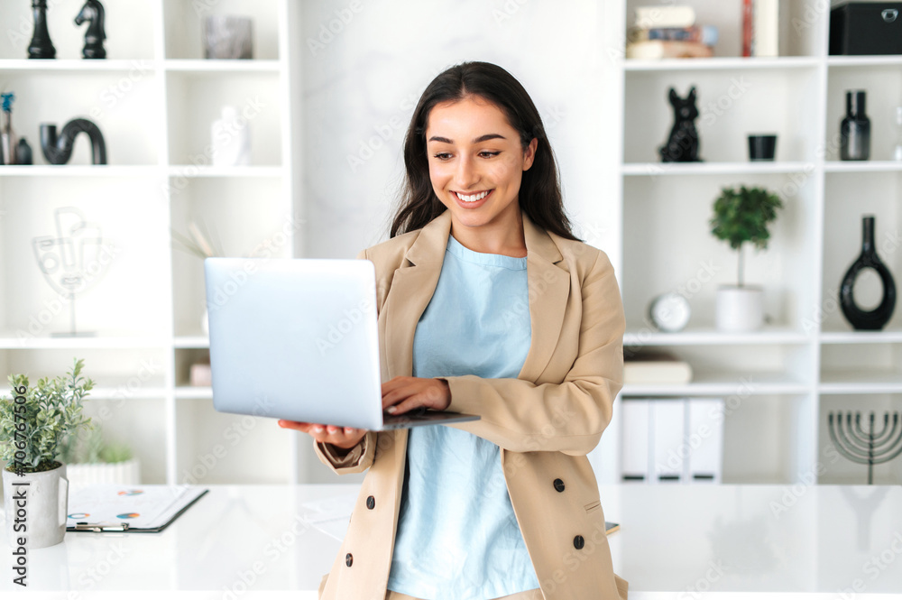 Successful positive brunette arabian or indian woman, business lady, company ceo, dressed in elegant suit, stand in a modern office near the table and holds an open laptop, looks at screen and smiling Successful positive brunette arabian or indian woman, business lady, company ceo, dressed in elegant suit, stand in a modern office near the table and holds an open laptop, looks at screen and smiling