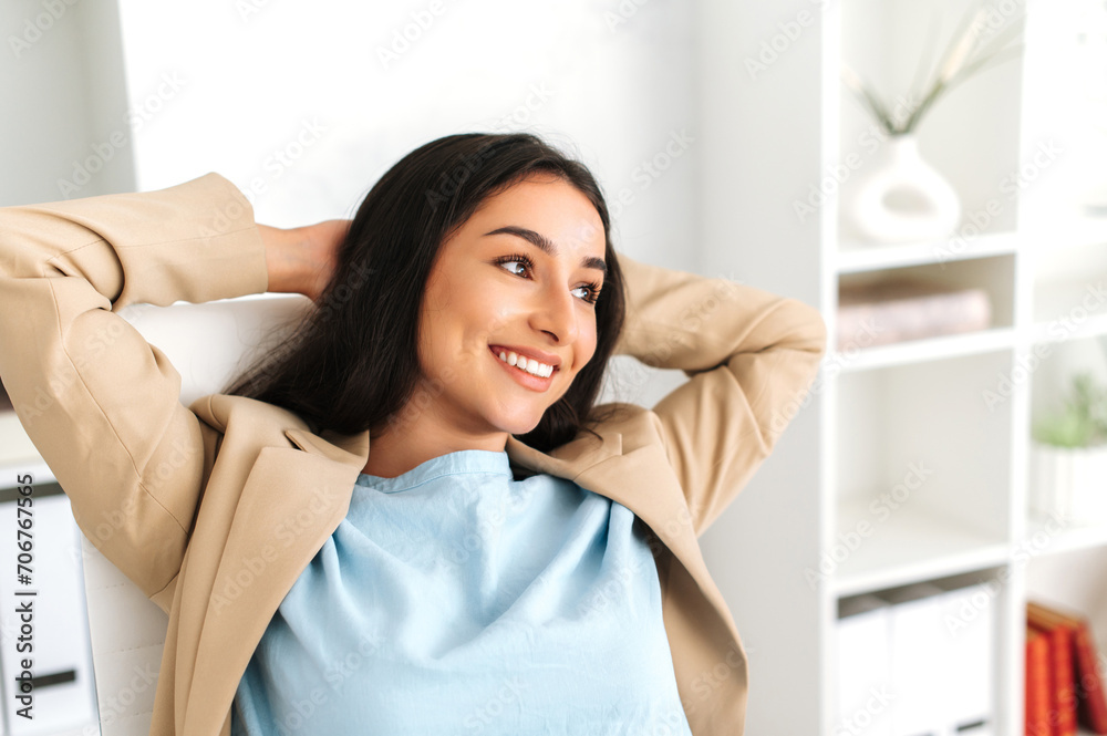 Happy satisfied carefree woman of indian or arabian nationality, sitting relaxed in a chair at her desk with her hands behind her head, looks away, taking a break from work tasks, smiling, dreaming Happy satisfied carefree woman of indian or arabian nationality, sitting relaxed in a chair at her desk with her hands behind her head, looks away, taking a break from work tasks, smiling, dreaming