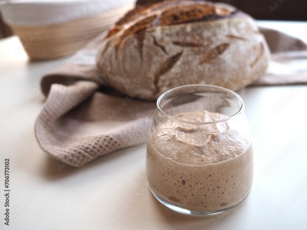 Closeup on fresh sourdough in a glass jar, bread and banneton in background, white table Closeup on fresh sourdough in a glass jar, bread and banneton in background, white table