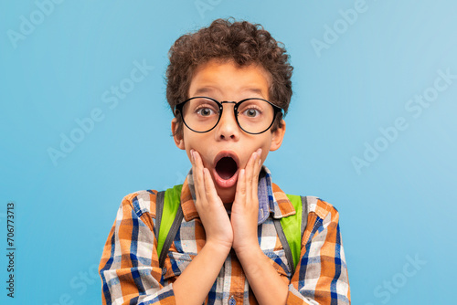 Shocked boy with glasses, hands on cheeks, blue background photo