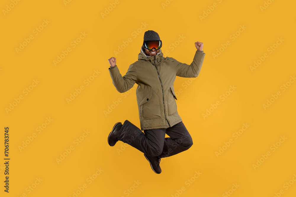 Winter sports. Happy man in ski suit and goggles jumping on orange background
