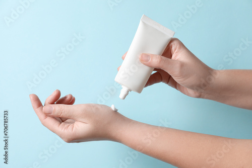 Woman applying cosmetic cream from tube onto her hand on light blue background, closeup