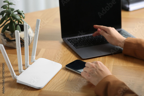 Woman with smartphone and laptop connecting to internet via Wi-Fi router at table indoors, closeup