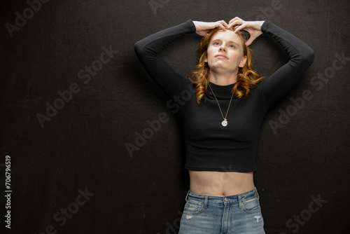 Young woman with red hair in black sweater and jeans poses in studio before a black background