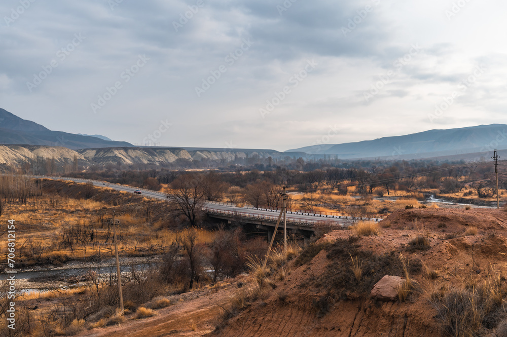Fototapeta premium Panorama of a mountain valley, road and bridge over a river in a mountainous countryside. Brown earth and bald trees.