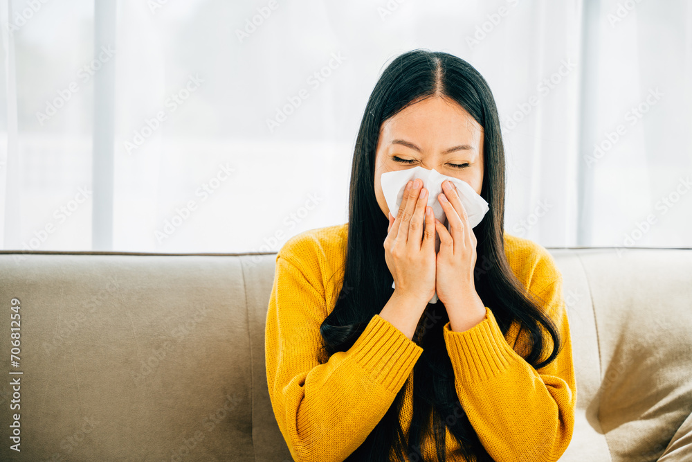 In an apartment an Asian woman holds a tissue blowing her nose due to a stuffy nose and coughing. Portrait of a patient at home seeking relief from illness. Snot and handkerchief in hand. In an apartment an Asian woman holds a tissue blowing her nose due to a stuffy nose and coughing. Portrait of a patient at home seeking relief from illness. Snot and handkerchief in hand.