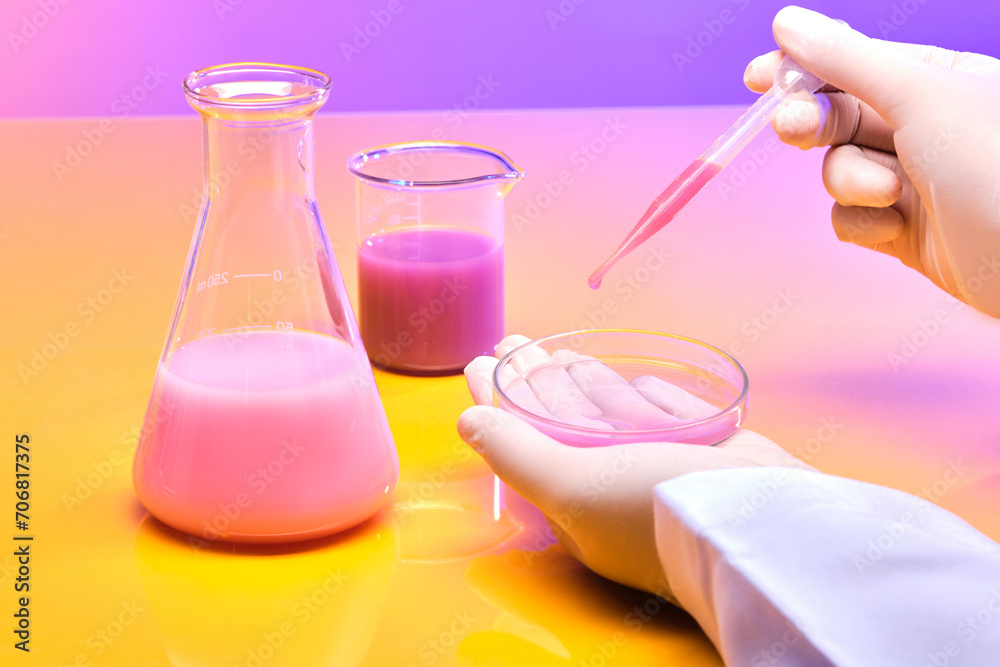 A hand holds a dropper containing pink liquid, next to an erlenmeyer flask and a beaker on a yellow pink gradient background. Product research and development. A hand holds a dropper containing pink liquid, next to an erlenmeyer flask and a beaker on a yellow pink gradient background. Product research and development.