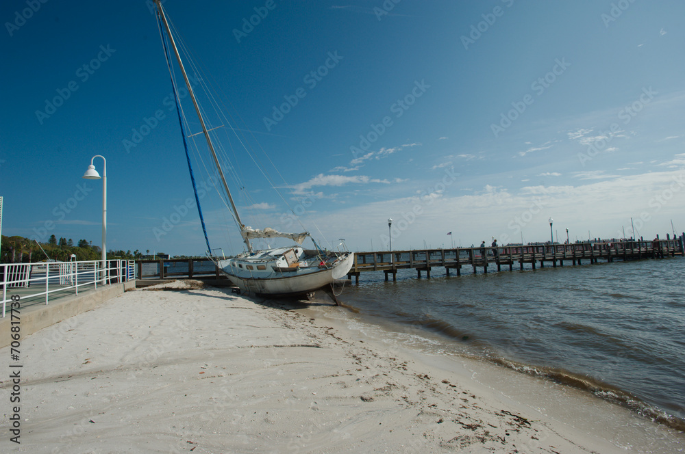 White and light blue sailboat with mast grounded on the beach by pier bridge in Gulfport, Florida after a storm. Sand in front leading line to boat and lamp post on the left. Sunny day with blue sky.
