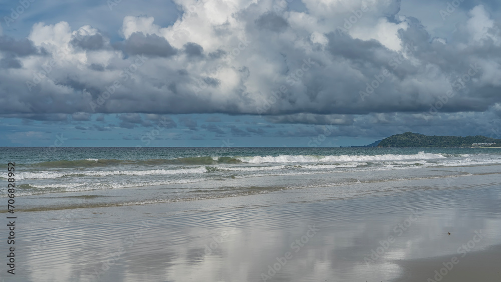 The waves of the turquoise ocean roll towards the shore, foaming and spreading along the beach. Clouds and blue sky are reflected on the wet smooth sand. A hill in the distance. Malaysia. Borneo. 