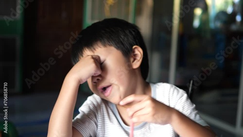 Asian child boy feel sleepy and tired, rubbing eyes, and yawn with exhausted, bored face while doing school homework indoor at home. Unhappy young kid concept.