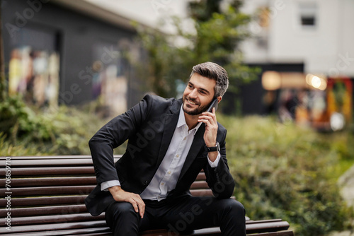 A happy businessman is sitting on the bench in park and having phone conversation.