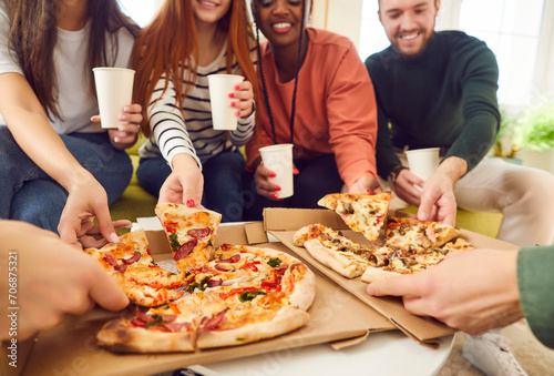 Happy smiling young people friends gathering in pizzeria or at home together eating tasty Italian food taking pizza slices from box enjoying party with cardboard cups in hands. Food delivery concept. photo