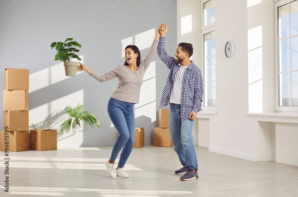 Happy young couple celebrating moving day with cardboard boxes in background smiling and dancing with plant in hands in new appartment. Family having fun. Relocating, real estate, mortgage concept. Happy young couple celebrating moving day with cardboard boxes in background smiling and dancing with plant in hands in new appartment. Family having fun. Relocating, real estate, mortgage concept.