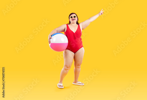Funny young happy fat woman in red swimsuit having fun holding inflatable ball and going on summer holiday trip standing on yellow studio background. Vacation tour and travel concept.