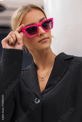 Stylish beautiful young glam woman model in a fashion black blazer puts on pink sunglasses indoors