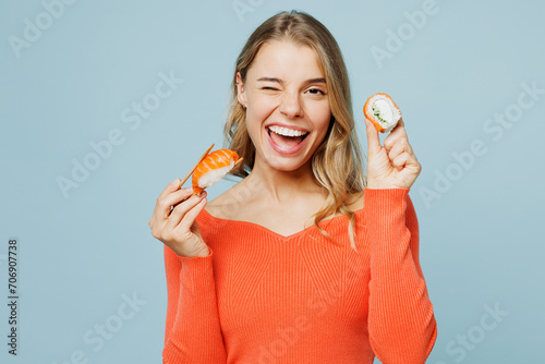 Close up young happy woman wear orange casual clothes look camera hold eat raw fresh sushi roll Japanese food with chopsticks wink isolated on plain blue background studio portrait. Lifestyle concept photo